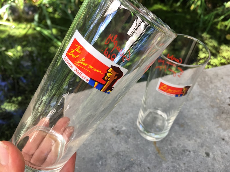May include: Two clear glass drinking glasses with red and blue labels that say "The  But Barman Johnnie Walker" and feature a cartoon illustration of a man with a mustache.