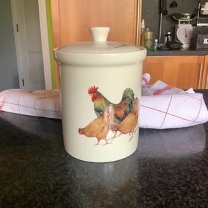 Peut inclure: Un pot en céramique crème avec un couvercle, orné d'une illustration de coq et de poules. Le pot est cylindrique avec une petite poignée sur le couvercle. Le coq est rouge, vert et jaune, et les poules sont brunes.