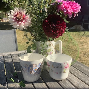 Op de afbeelding: Twee witte keramische bekers met bloemenmotieven staan op een houten tafel. Een beker heeft het woord "Seecile" erop geschreven. De bekers worden omringd door een boeket roze, rode en witte bloemen.