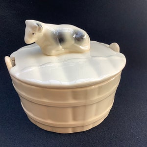 May include: A white ceramic butter dish shaped like a barrel with a lid featuring a black and white cow figurine. The dish has a cream-colored glaze.