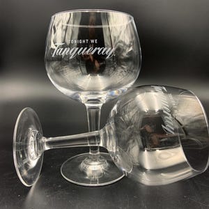 May include: Two clear, stemmed gin glasses with a rounded bowl and a wide base. The text "TONIGHT WE Tanqueray" is printed in white on the bowl of each glass. One glass is upright, and the other is lying on its side.