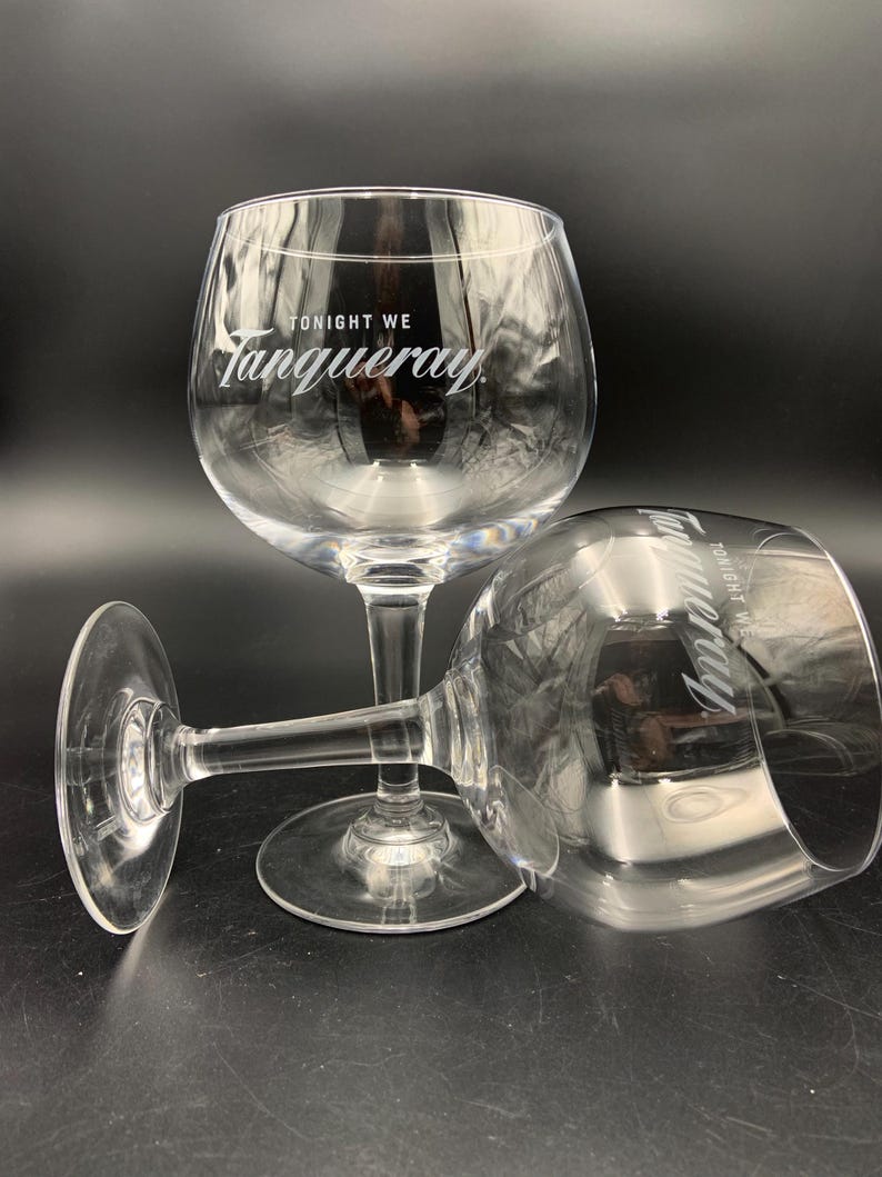 May include: Two clear, stemmed gin glasses with a rounded bowl and a wide base. The text "TONIGHT WE Tanqueray" is printed in white on the bowl of each glass. One glass is upright, and the other is lying on its side.
