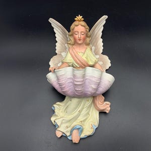 Puede incluir: Figura de ángel de cerámica con corona dorada y grandes alas. El ángel sostiene un cuenco grande en forma de concha con un degradado rosa y morado. El ángel lleva un vestido amarillo con detalles azules.