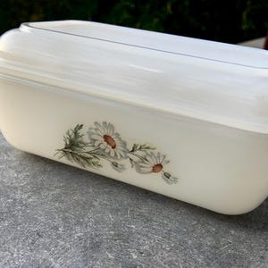 Puede incluir: Una fuente rectangular de color blanco roto con tapa transparente. La fuente presenta un diseño floral de margaritas blancas con centros amarillos y hojas verdes. La fuente está sobre una superficie gris.