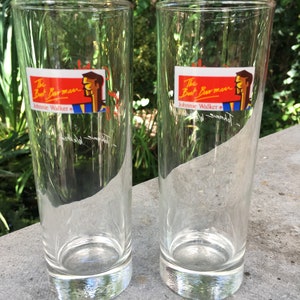 May include: Two clear glass highball glasses with a red and yellow label that reads "The Best Barman Johnnie Walker" with a cartoon illustration of a man.