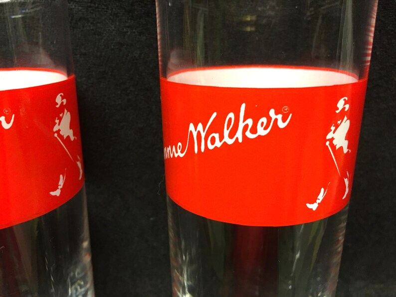 May include: Two clear glass tumblers with a red band around the top. The band has the words "Johnnie Walker" in white lettering and a white silhouette of a man walking with a cane.