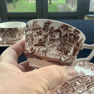 May include: A set of two brown and white teacups and saucers featuring a country cottage scene design. The cups have a handle and the saucers have a scalloped edge.