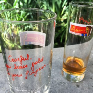 May include: Two clear glass tumblers, one with a red handwritten message that reads "Careful.  Gold you leave good is your fingers." and a signature at the bottom, and the other with a sticker that reads "The Barman" and a cartoon image of a man.