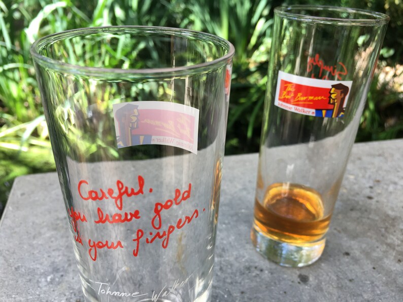 May include: Two clear glass tumblers, one with a red handwritten message that reads "Careful.  Gold you leave good is your fingers." and a signature at the bottom, and the other with a sticker that reads "The Barman" and a cartoon image of a man.