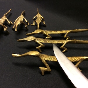 May include: A set of six gold-coloured metal lizard-shaped knife rests with a silver knife on a black surface.