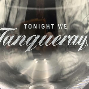 May include: Clear glass stemware with the words "TONIGHT WE" and "Tanqueray" in white lettering. The glass has a rounded bowl and a long stem, suitable for cocktails.