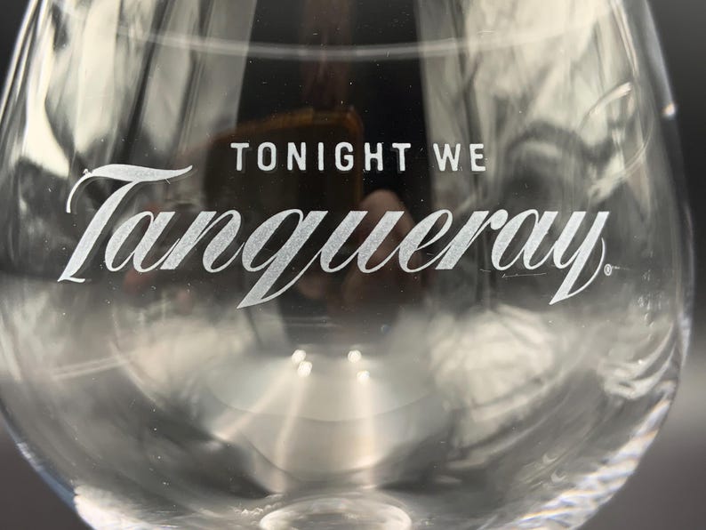 May include: Clear glass stemware with the words "TONIGHT WE" and "Tanqueray" in white lettering. The glass has a rounded bowl and a long stem, suitable for cocktails.