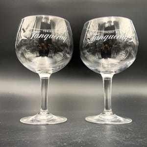 May include: Two clear, stemmed gin glasses with a rounded bowl. The text "TONIGHT WE Tanqueray" is printed in white on the bowl of each glass. The glasses are on a dark surface.