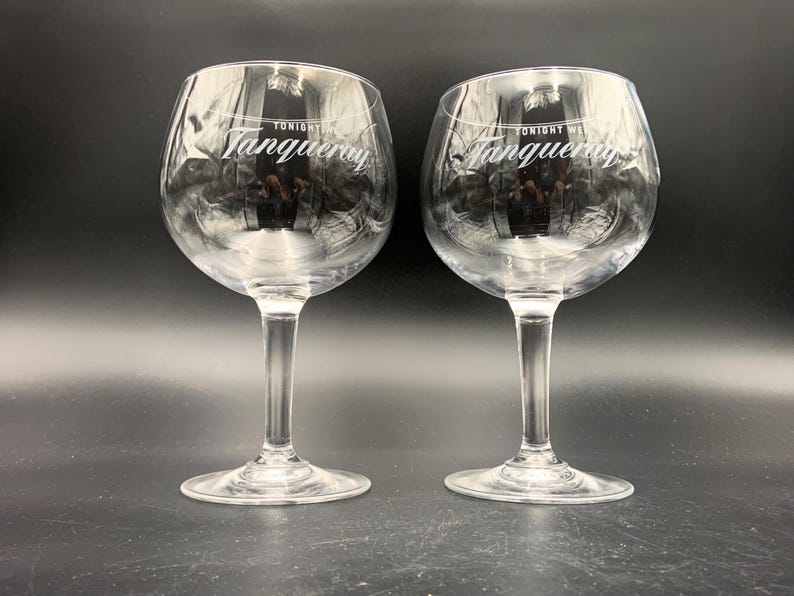 May include: Two clear, stemmed gin glasses with a rounded bowl. The text "TONIGHT WE Tanqueray" is printed in white on the bowl of each glass. The glasses are on a dark surface.