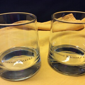 May include: Two clear glass tumblers with the words "Johnnie Walker" printed on the bottom. The glasses are sitting on a yellow surface.