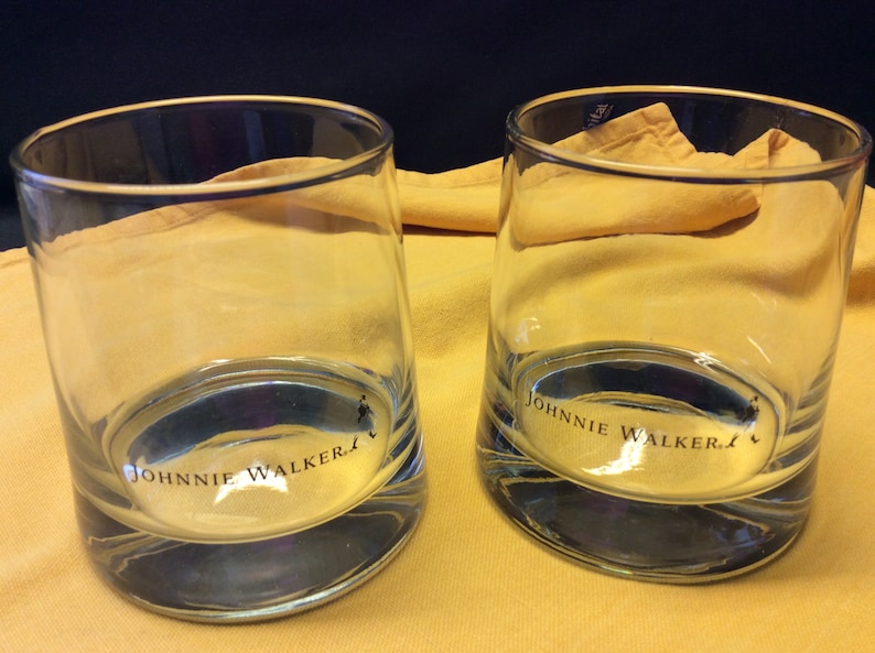 May include: Two clear glass tumblers with the words "Johnnie Walker" printed on the bottom. The glasses are sitting on a yellow surface.