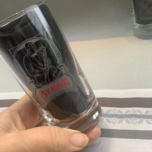 May include: Clear glass tumbler with a black interior. The glass features a vintage design with a saxophone player and the word "BYRRH" in red lettering. The glass is held at an angle.