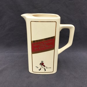 Pode incluir: Jarro de cer&acirc;mica creme, com formato retangular e al&ccedil;a. Apresenta um r&oacute;tulo vermelho e dourado com o texto "Johnnie Walker Old Scotch Whisky". Uma pequena ilustra&ccedil;&atilde;o de um homem a andar est&aacute; abaixo.