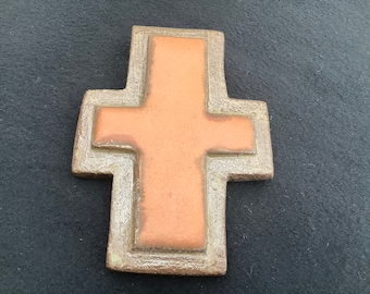 May include: A terracotta cross with a raised, textured border. The cross is a light brown color and has a rustic, handmade look.