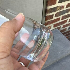 Pu&ograve; includere: Un bicchiere trasparente e curvo con fondo arrotondato. Il bicchiere &egrave; tenuto in mano, con la luce che si riflette sulla superficie liscia. Lo sfondo comprende un muro di mattoni e una superficie grigia.