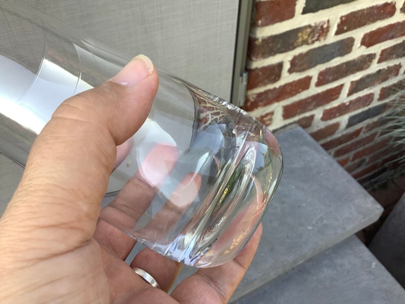 Pu&ograve; includere: Un bicchiere trasparente e curvo con fondo arrotondato. Il bicchiere &egrave; tenuto in mano, con la luce che si riflette sulla superficie liscia. Lo sfondo comprende un muro di mattoni e una superficie grigia.