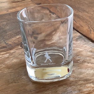 May include: A clear glass tumbler with a square base and a walking man logo etched on the bottom. The logo says "Johnnie Walker".