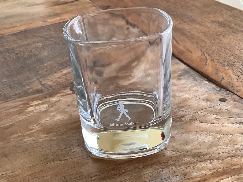 May include: A clear glass tumbler with a square base and a walking man logo etched on the bottom. The logo says "Johnnie Walker".