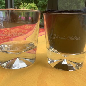 May include: Two clear, lowball whiskey glasses with a geometric base. One glass is empty, the other contains a dark substance and a black napkin. The text "Johnnie Walker" is visible on the glass.