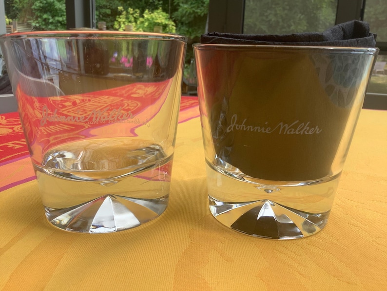May include: Two clear, lowball whiskey glasses with a geometric base. One glass is empty, the other contains a dark substance and a black napkin. The text "Johnnie Walker" is visible on the glass.