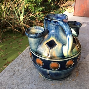 May include: A blue and brown ceramic vase with three openings and a handle. The vase has a geometric design with a diamond shape and circular patterns.