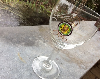 May include: A clear glass beer glass with a gold inscription and a yellow, green, and red logo with the text "Opifex" on it.
