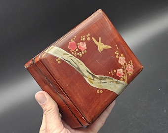 Può includere: Una scatola quadrata di legno con finitura marrone rossiccio lucida. Il coperchio è decorato con un uccello dorato, fiori rosa e un disegno dipinto bianco e oro. La scatola è tenuta in mano da una persona.