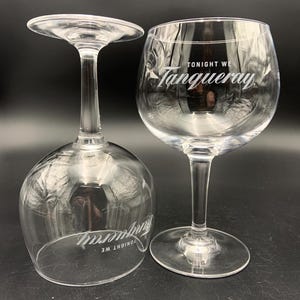 May include: Two clear, stemmed gin glasses. One glass is upright, the other is upside down. The text "TONIGHT WE Tanqueray" is printed on the glasses in white. The glasses are on a black surface.