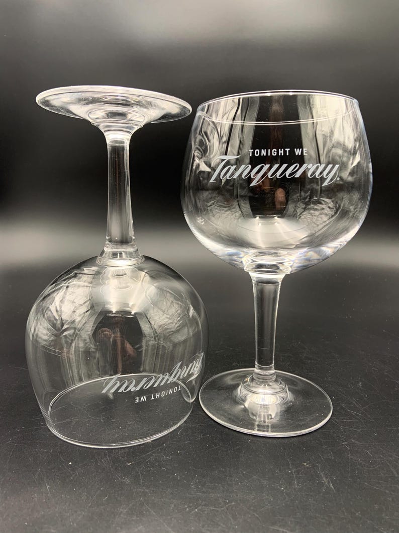 May include: Two clear, stemmed gin glasses. One glass is upright, the other is upside down. The text "TONIGHT WE Tanqueray" is printed on the glasses in white. The glasses are on a black surface.