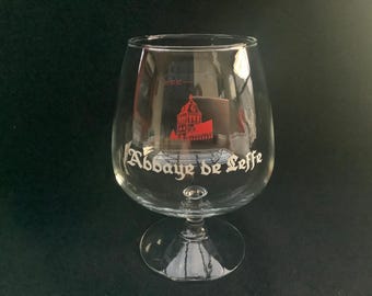 May include: A clear, stemmed beer glass with a rounded bowl and a wide base. The glass features the words "Abbaye de Leffe" in white script, along with a red illustration of a building. The glass is designed for enjoying beer.