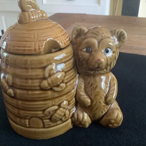 May include: A ceramic honey pot shaped like a beehive with a bear figurine on the side. The pot is brown and has bees and honeycomb details. The bear is brown and has a white nose and eyes.
