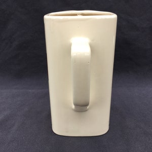 Pode incluir: Uma caneca retangular de cer&acirc;mica creme com uma al&ccedil;a. A caneca tem um acabamento liso e brilhante e um design simples e minimalista. A al&ccedil;a &eacute; fixada na lateral da caneca. A caneca tem aproximadamente 15 cm de altura.