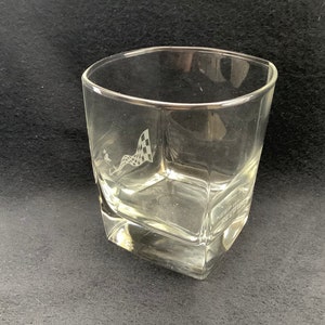 May include: A clear glass tumbler with a chequered flag design etched on the side. The glass has a square shape with rounded edges.