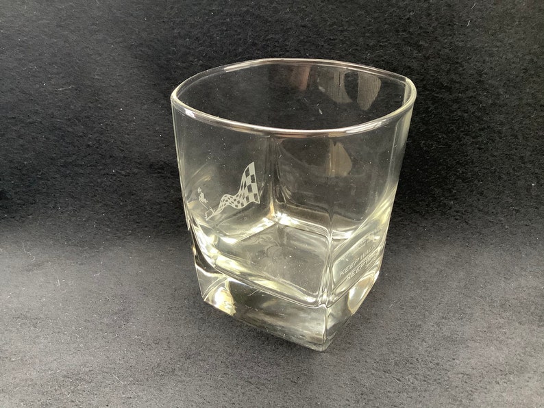 May include: A clear glass tumbler with a chequered flag design etched on the side. The glass has a square shape with rounded edges.