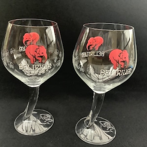 Puede incluir: Dos vasos de cerveza de vidrio transparente con tallo curvo. Cada vaso presenta un elefante rosa con el texto "Delirium" y "Tremens" impreso en el vaso.