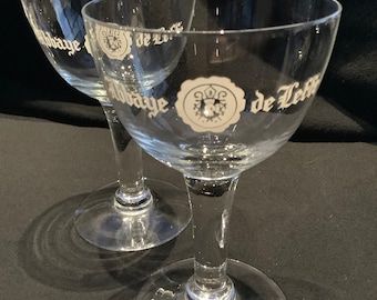 May include: Two clear stemmed glasses with a wide bowl. Each glass has a white emblem and text that reads "Abbaye de Leffe". The glasses are on a dark surface.