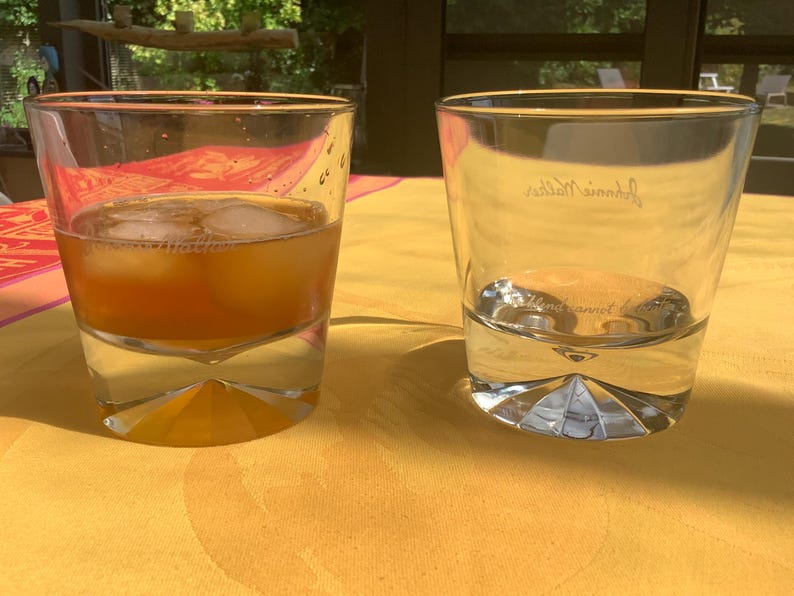 May include: Two clear, lowball whiskey glasses on a yellow tablecloth. One glass contains an amber-colored liquid with ice cubes. The other glass is empty with the brand name etched on the side. The glasses have a geometric base.