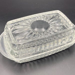 May include: Clear glass butter dish with a rectangular shape and a decorative, textured pattern. The lid features a central circular indentation and a floral-like design. The dish has a curved base and a handle.