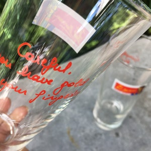 May include: Two clear glass drinking glasses. One glass has the words "Careful. You have got your fingers" written in red marker.