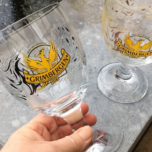 May include: Two clear Grimbergen beer glasses. The glasses have a stem and a wide bowl. The Grimbergen logo, featuring a yellow phoenix, is printed on the front. The text "Grimbergen" and "Bieres d'Abbaye Anno 1128" are also visible.