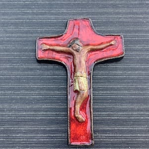 May include: A decorative cross featuring a figure of Jesus Christ. The cross is red with a brown border, and the figure is depicted in brown and gold tones. The background is a dark gray, textured surface.