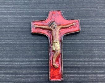 May include: A decorative cross featuring a figure of Jesus Christ. The cross is red with a brown border, and the figure is depicted in brown and gold tones. The background is a dark gray, textured surface.