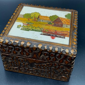 May include: A vintage wooden box with a detailed carved exterior. The lid features a painted landscape scene with a river, houses, and a boat. The box is rectangular and has a dark brown finish.