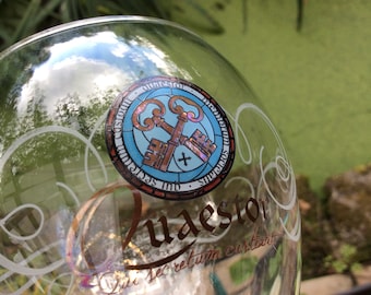 May include: Clear glass with a decorative emblem featuring two crossed keys in a stained-glass style, surrounded by text. The glass also has etched swirls and text. The image is taken outdoors.