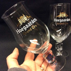 Op de afbeelding: Twee heldere Hoegaarden Grand Cru bierglazen. De glazen hebben een gebogen vorm en een steel met een decoratieve voet. Het Hoegaarden-logo is in wit en goud op het glas gedrukt.